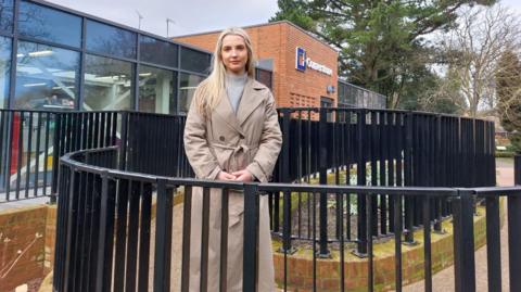 A blonde haired woman in a beige coat and grey roll neck jumper stands behind a black metal fence smiling at the camera. 