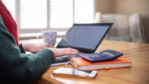A picture of someone typing on a keyboard. They are wearing a green jumper and a red scarf. There is a book, phone and calculator on the table next to the laptop. There is a mug in the distance.