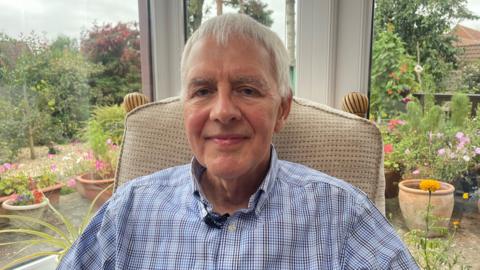 A man in his 60s with short white hair, looking towards the camera. He is wearing a light blue check shirt. He is sitting in a beige chair in his conservatory, with a view of his garden behind him. There are lots of plants and flowers on show in the garden.