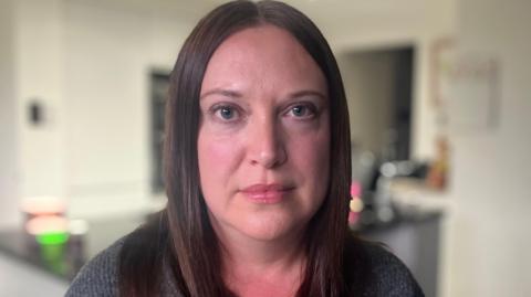A woman with dark hair looks at the camera in a kitchen. She wears a grey jumper