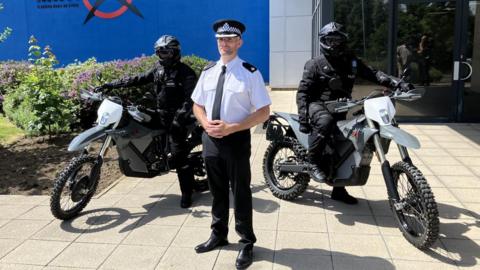 Supt Paul Richardson, wearing a uniform of a white short-sleeved shirt with police patches, black tie and trousers and a police officer's cap, standing between two riders on the new, lightweight electric motorbikes which have a similar shape and design to motocross bikes. The riders themselves wear all black, including blacked out helmets.