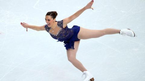 Jenna McCorkell has her brown hair up bun. Her arms are outstretched and she is skating on one skate. She is wearing a dark blue ice-dancing dress which is covered in sequins. 