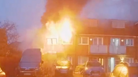 A house on fire with roaring flames coming out of windows and dark smoke in the sky. There are two cars and one van in front of the house.