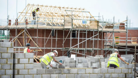 Builders on a housing site. In the foreground three builders cement bricks together. In the background a builder stands on top of a mostly built red brick house, with the exposed wooden roof beams also in place. 