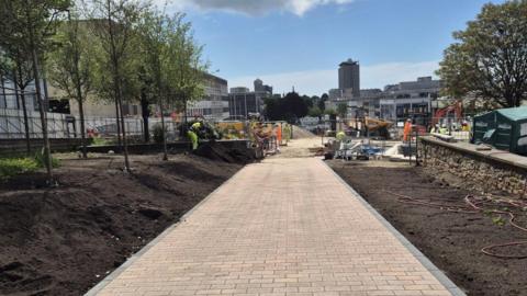Granite paving along Armada Way. Soil lines either side of the pathway. A worker, dressed in a hi-vis tabard, is stood on the soil. Trees have been planted. Machinery can be seen on the site. Plymouth's Civic Centre is in the distance.