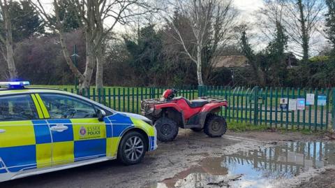 A marked blue and yellow Essex Police car is parked on a muddy road. A red and black quad bike is parked in front of it. Behind the bike are green metal railings that look like they lead to a park.