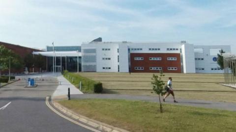 A man with a backpack walking in front of the Ordnance Survey building on a sunny day. The building is white with red brick areas. A well maintained lawn is laid out alongside the entrance road.