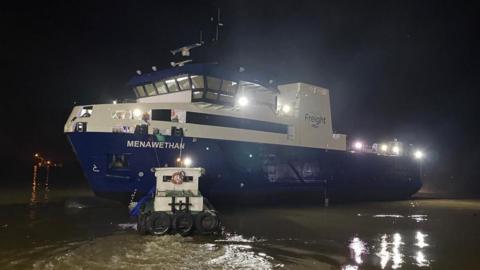 A navy blue a white ship in water on a dark evening, with the name of the ship 'Menawethan' and the word 'Freight' on the side.