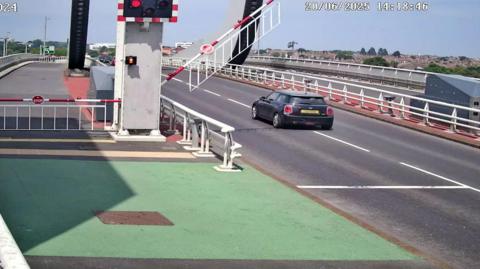 A traffic camera image of a Mini Cooper car passing red safety lights on the Gull Wing Bridge. A barrier is lowering behind the car.