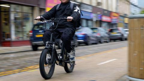 A man on a bike wearing all black. The background is slightly blurred with cars parked along the side of the pavement visible.