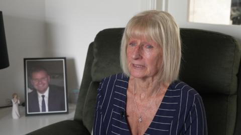 Carol Coleman, who has blonde hair and blue eyes. She is sat in a greem armchair wearing a blue dress with white pinstripes. A photo of Dean, who is smiling and wearing a black suit, is in the foreground.