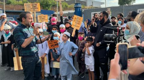 A man holding a small megaphone addresses a gathered crowd of men, women and children. Some of them wear face masks or hold their hands over their mouths and noses. Others hold home made placards saying things like 'end the stink' and 'stop toxic fumes'.
