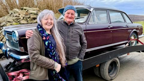 A woman with long grey hair stands next to a man with a blue woollen hat. They are standing in front of an old brown car, which is on the back of a lorry.