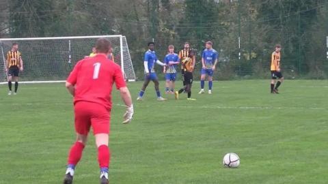 A scene from a football match with a goalkeeper in a red kit and with number 1 on his back readying himself to take a free kick from inside his own half.