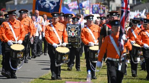 A group of people dressed in orange uniforms, wearing caps and carrying drums march in rows in Cowdenbeath in 2018.