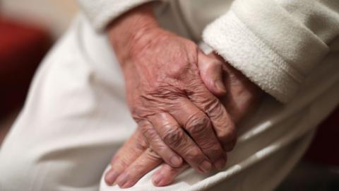 The hands of an elderly woman placed on top of each other. She is wearing a white fleece and trousers.