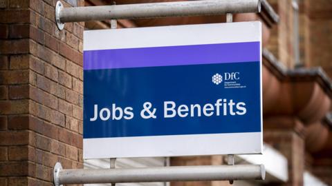 Sign outside benefits office in Belfast. The sign says Jobs & Benefits in white, with a blue and purple background. It also has the department for communities sign on the right hand side. 