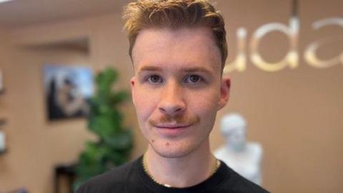 Hair salon owner, Sam Cowan, is staring into the camera. He has short light brown hair and a moustache. He's wearing a black t-shirt with a gold necklace and he's standing in his hair salon. Behind him is the signage for his business, which is called Audacity, along with a large green plant and a white roman-style decorative bust.