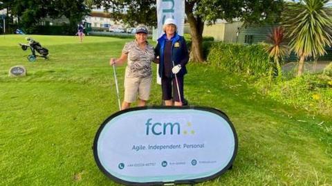 A photo which shows two women wearing hats and holding golf clubs. The woman on the left is wearing a patterned beige and black top and beige long shorts. The woman on the right is wearing a blue and black jacket, a black top and black trousers. They are on a golf course which has green grass surrounding them. In front of them is a sign that says FCM agile, independent, personal.