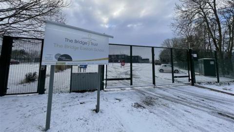 The Bridge Easton School front gates surrounded by snow. The school sign is on two poles in front of a high metal fence and gate, which is shut. A large car park beyond the gate is white with ice and snow. Only one car is parked.