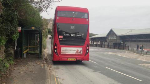 A red double-decker bus pulls away from a green bus stop shelter in Padstow.