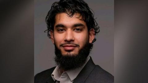Adam Khan is sitting, looking at the camera and smiling. He has black hair and black facial hair. He is wearing a grey suit jacket and light grey shirt.
