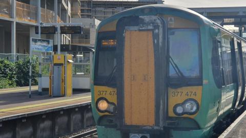 A Southern Train, in its green and gold livery, stands at a platform at East Croydon station, with new tower block construction in the background, on a sunny day.