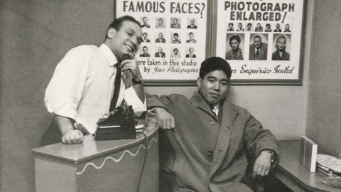 Dave Sharkey, with customer, 1962. Black‑and‑white photograph of Dave Sharkey, with customer. Dave leans against a counter with a camera placed on top, while the other sits beside the desk. Posters of sample portrait photographs hang on the wall behind them.