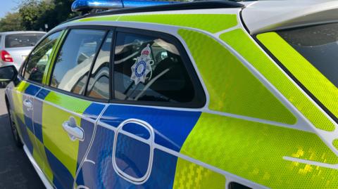 A close-up of the passenger side of a police car marked with blue and luminous yellow livery and a Humberside Police insignia in the rear side window.