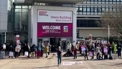 A group of people are standing outside Sheffield Hallam University with pink and white placards. In the background is a large entrance with a pink and white sign