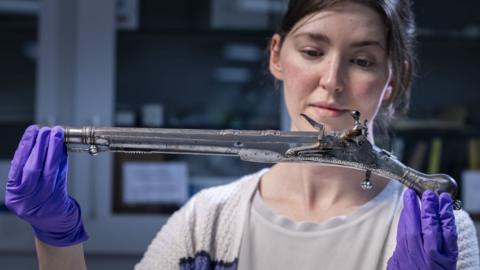 Curator Ella Paul holds one of the Jacobite pistols aloft. She is wearing a white top and white cardigan and purple protective rubber gloves.