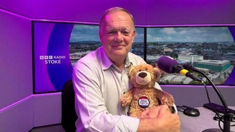 A man sitting in a radio studio with tv screens behind him is looking at the camera and holding a bear. The man is wearing a light-coloured shirt and has a purple microphone in front of him.