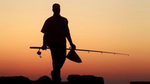 Silhouette of a man holding a bag in his right hand and a fishing rod in his left walking towards the sunset, rocks in front of him. Sun not in shot but the sky in front is ombre yellow, orange to dark brown.