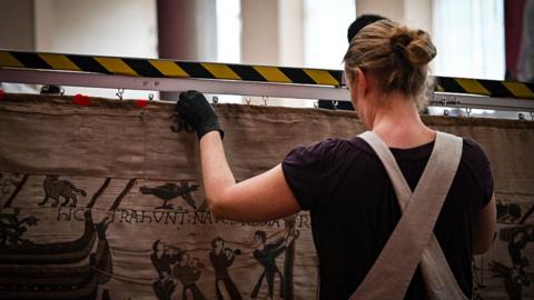 A photograph of a gallery worker carefully inspecting the Bayeux Tapestry. The worker is a woman and the photo is taken from behind. She is wearing overalls, has her hair tied back and has black gloves on her hand. 
