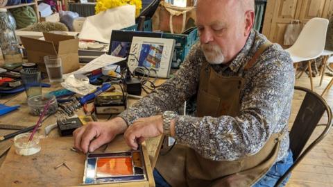 A man sites at a large table working on a rectangular piece of stained glass. The inside of the design is orange and is surrounded by four pieces of dark blue glass. He is wearing a brown apron and is looking down at his work.