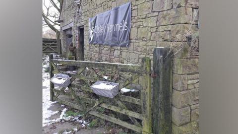 Sign on the wall of a grey stone building reads "Disley Kennels". There is snow on the ground of a paved are outside the rural-looking building. There is an open wooden gate.