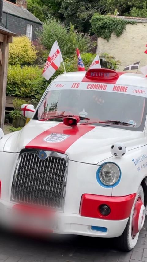 A white and red car which has "its coming home" written in red letters. There are english flags and footballs over the car. The number plate is blurred.