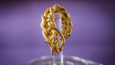 A close-up of the fragment of gold arm-ring. It has eight strands of gold rods which have been plaited together. It hangs on a clear stand in front of a purple background.