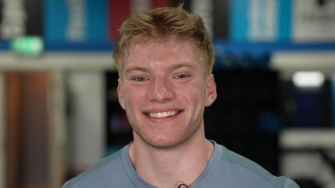 Blonde man in a blue t-shirt smiling at the camera