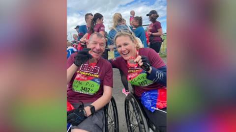 Stuart Hall and Claire Lomas in wheelchairs, both wearing burgundy tops, with medals around their necks after completing a challenge