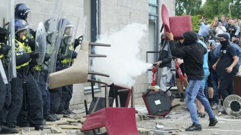 A man wearing blue tracksuit bottoms and a black hooded top throws a red chair at police officers.