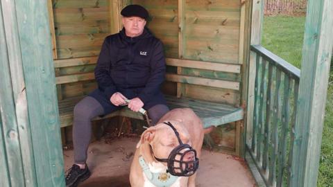 A man wearing a flat cap and a black jacket sitting on a green bench holding his XL Bully dog.