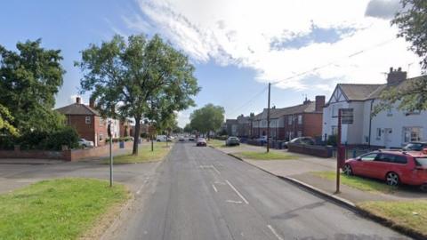 A google maps streetview image of Fryston Road. It is a fairly average residential street. Cars are driving up and down the road. The houses on either side of the street are quite large. There are cars parked near the houses, on driveways, and on the public grass verges outside. 