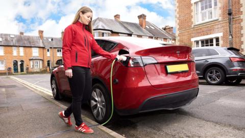 A red electric car parked on a residential street. A woman is standing on the pavement to the left of the car, plugging in a green electric charging cable. The cable runs along the pavement. The woman has long light brown hair, and is wearing black trousers and a bright red jacket. She has red and white trainers on.