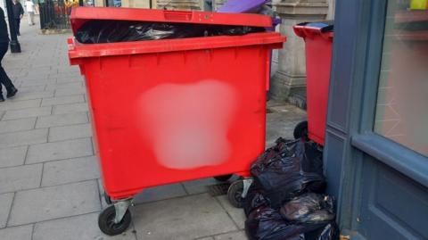 A large red Hills Waste bin left out on a street in Trowbridge, Wiltshire. A black bin bag is on the floor next to it. Two smaller wheelie bins can be seen next to the Hills bin. 