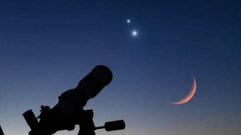 A close-up image of a silhouette of a telescope pointed up towards Venus and Jupiter in the sky.