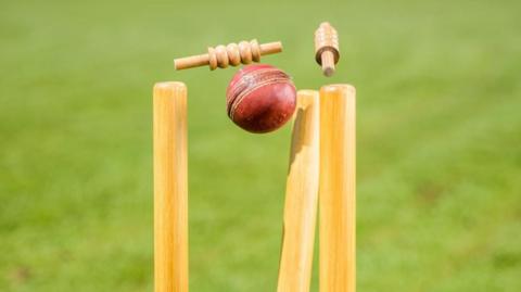 A cricket ball dislodging cricket stumps. The ball is red with a cream-coloured seam around the middle. The stumps are upright, wooden cylinders while the bails are smaller pieces of light wood.