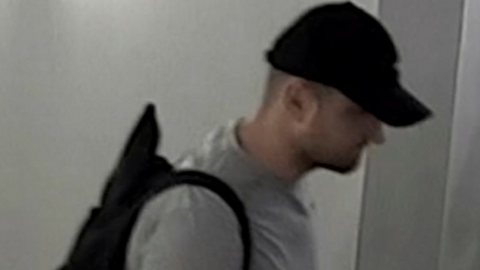 A low resolution CCTV image of a man in a grey t-shirt, dark coloured baseball cap, and dark coloured backpack. 
