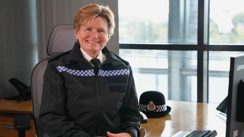 Maggie Blyth wearing a black police's uniform with blue and white checked stripe across the chest, with her police cap beside her. She is sitting at a desk in front of a computer with her elbows resting on the table. She has short blonde hair and is smiling at the camera. 