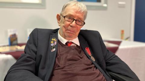 An elderly man sits down and smiles. He has short white hair, a suit jacket with a medal and badges, over a brown top over a white shirt and red tie.